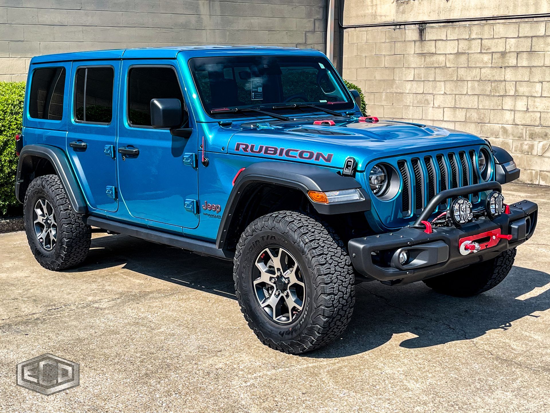 Blue Jeep Wrangler Rubicon parked beside a tan stone wall in bright sunlight