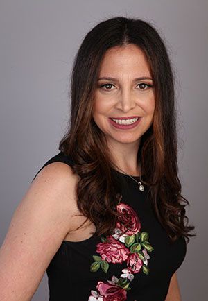 Smiling woman in a black dress with floral embroidery against a gray background