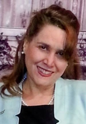 Smiling woman in a light blue blazer and pearl necklace against a purple curtain backdrop