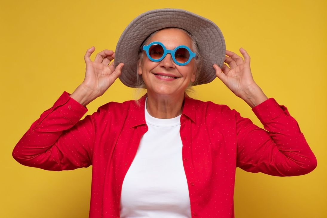 Smiling person in red jacket and blue sunglasses, adjusting a wide-brimmed hat against a yellow background
