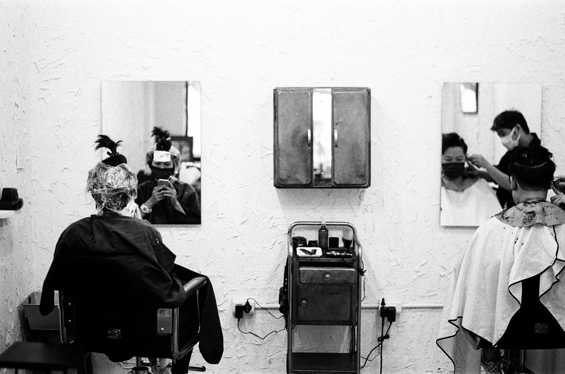 Interior of a hair salon; two clients and a stylist visible. Mirrors, equipment, and a cabinet are also present.