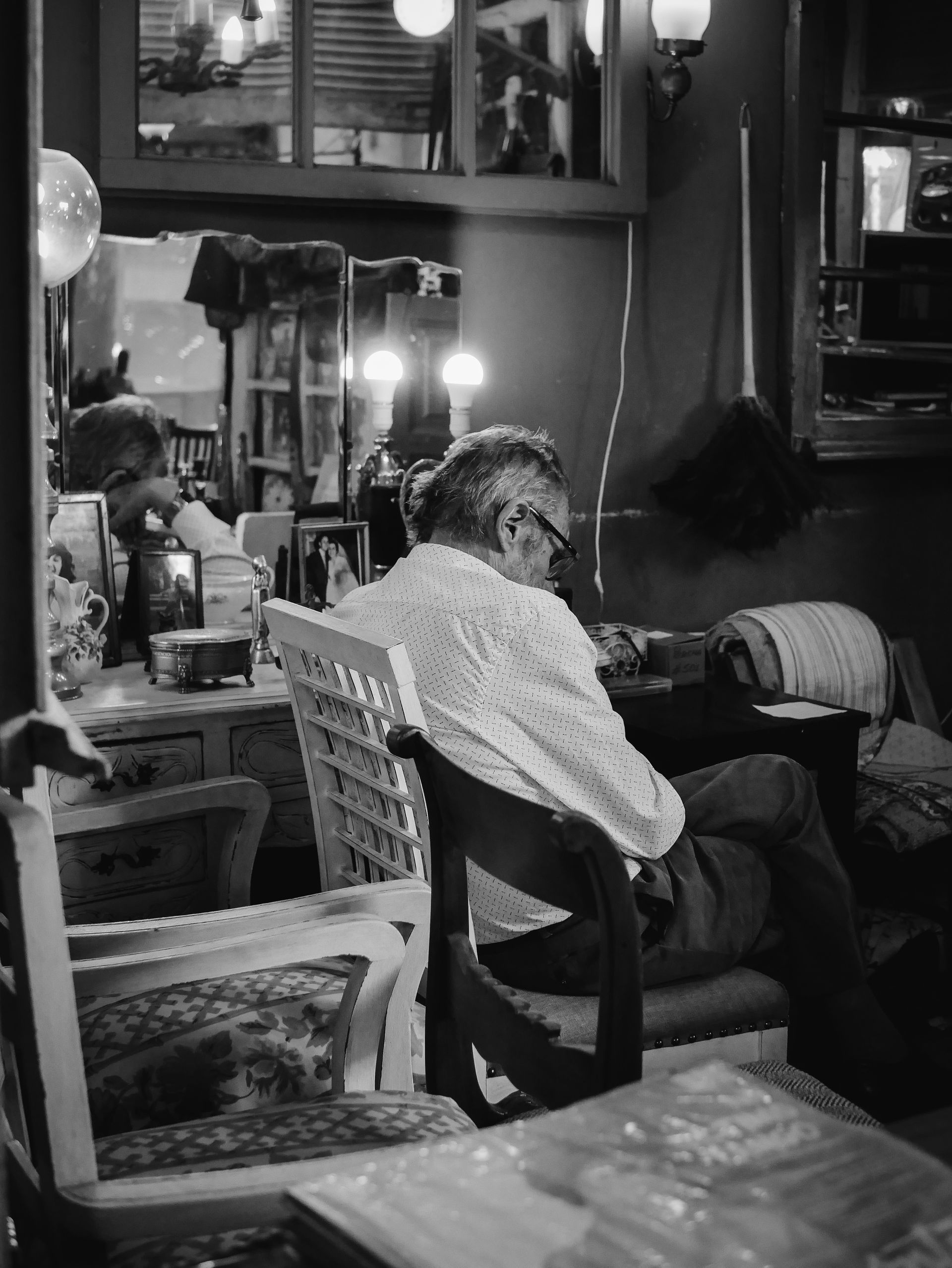 Man sits in a chair indoors, back to the camera. Dimly lit room with vintage furniture and decor.