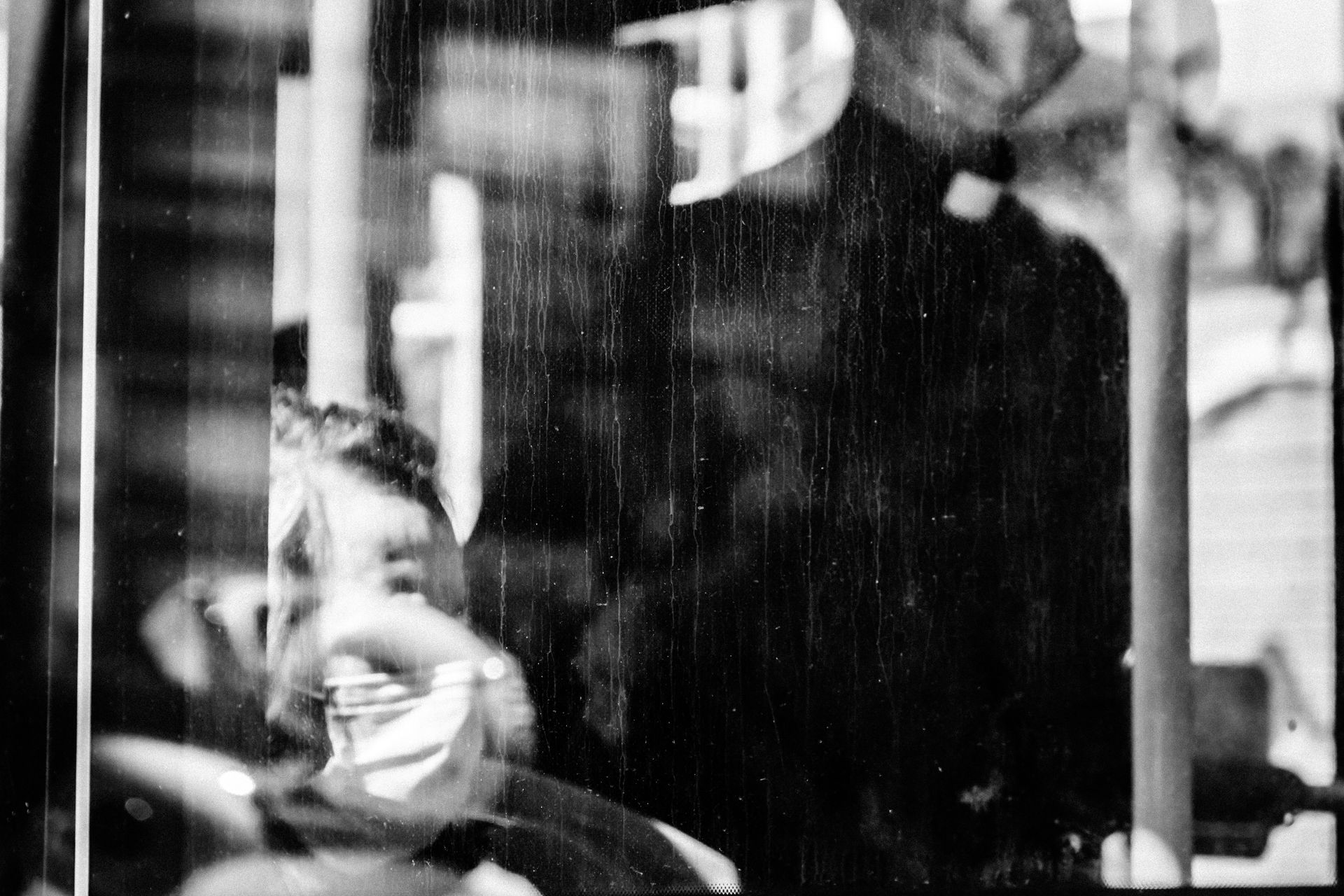 Man with surgical mask reflected in glass, obscured by streaks. Another person in dark clothes stands behind him.