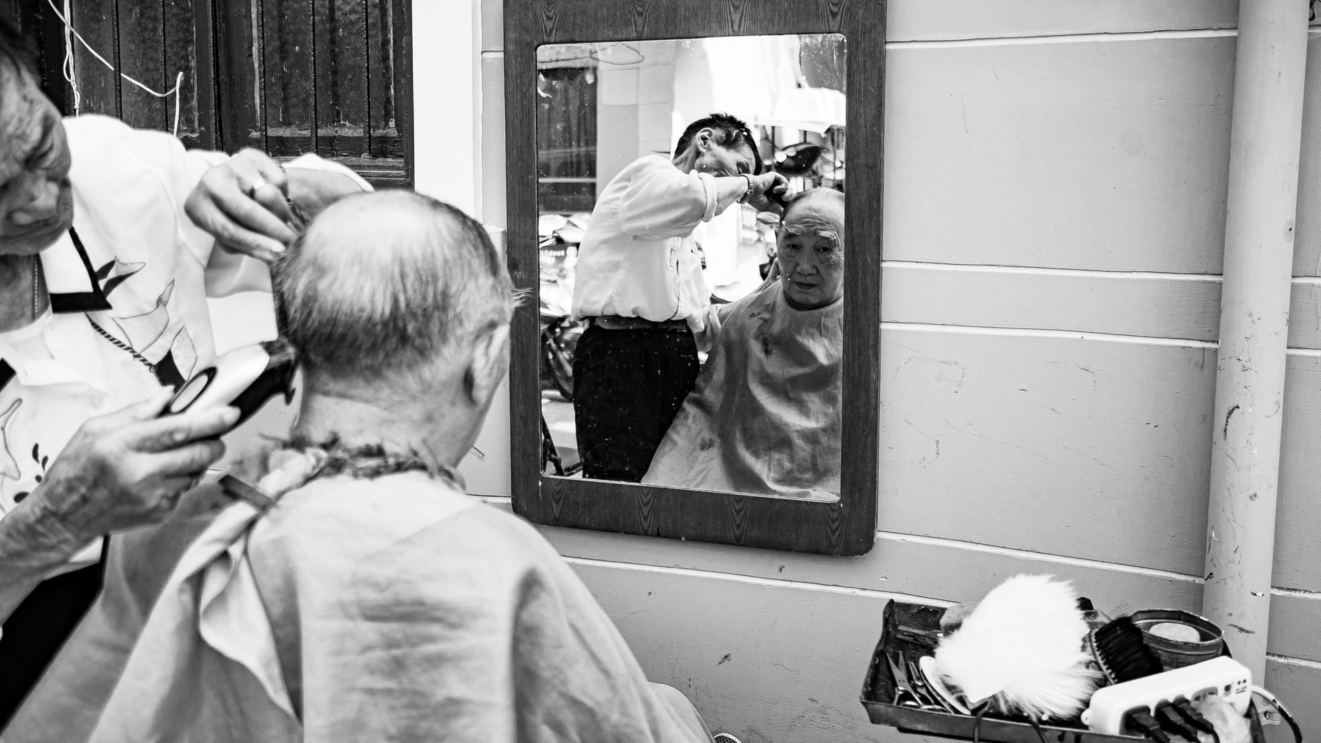 Barber cutting a person's hair outdoors, visible in mirror. Hair cutting tools and supplies are nearby.