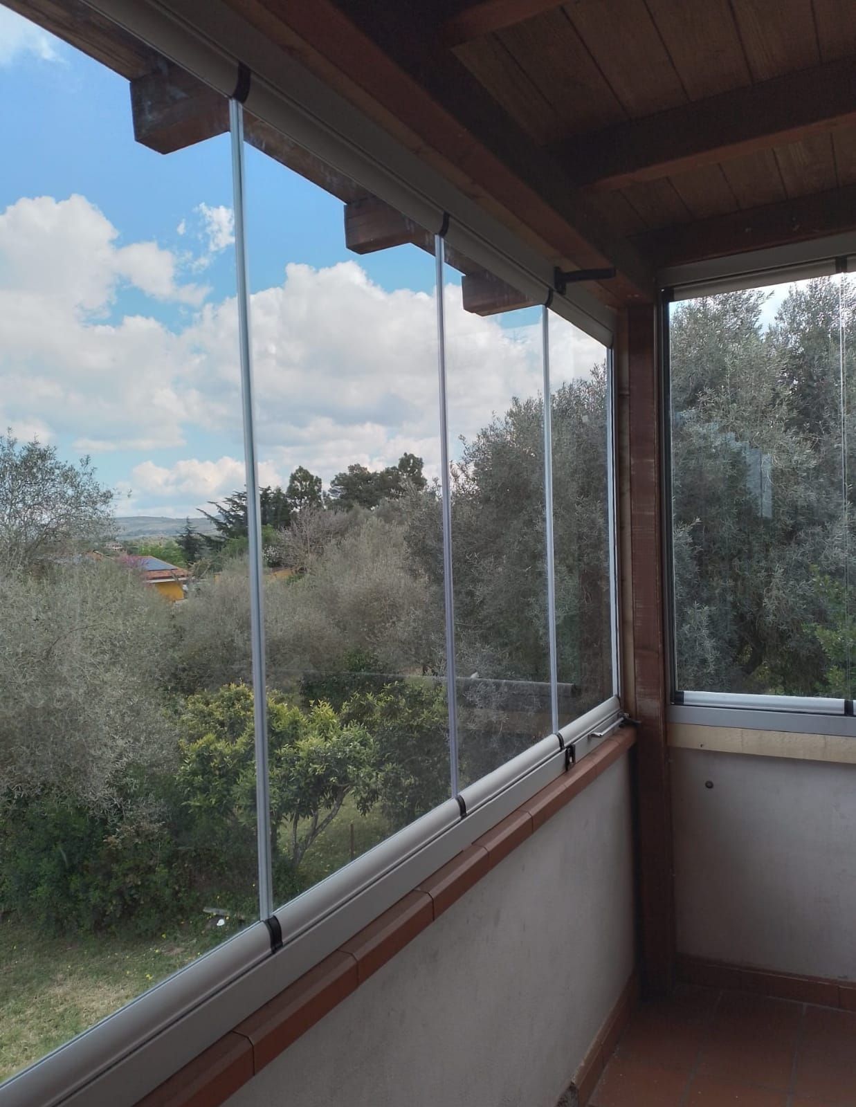 Balcone con pannelli in vetro e travi in legno, con vista su un paesaggio verde e cielo azzurro.