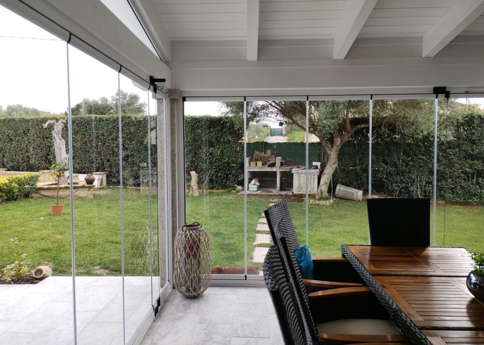 Patio con pareti in vetro che si affaccia su un prato verde con tavolo da pranzo e vegetazione da giardino.