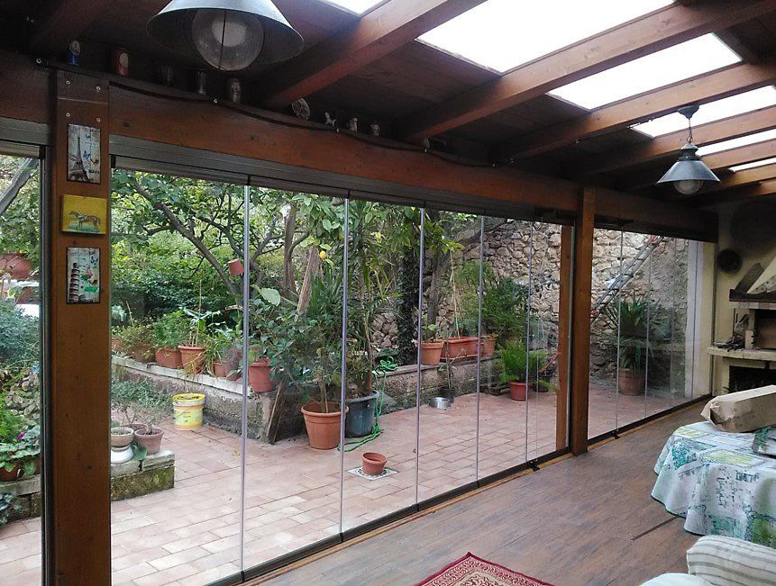 Veranda con pareti in vetro che si affacciano su un giardino con piante in vaso e un muro in pietra.