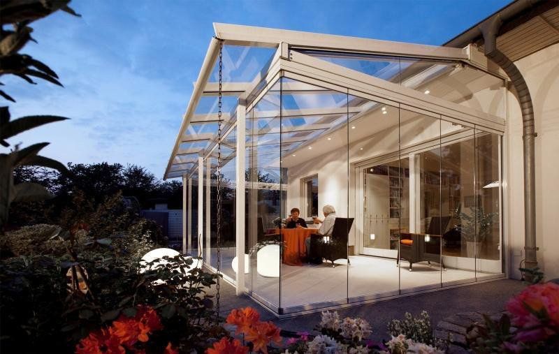 Veranda con vetrate annessa a un edificio, con una persona all'interno seduta a un tavolo. Il tutto circondato da un giardino.