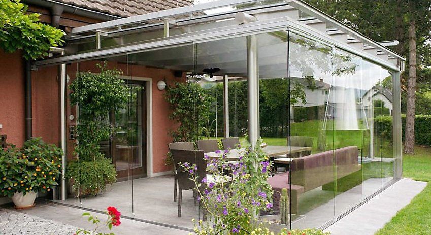 Patio con pareti in vetro, tavolo, sedie e una panca, accanto a una casa e a un cortile verde.