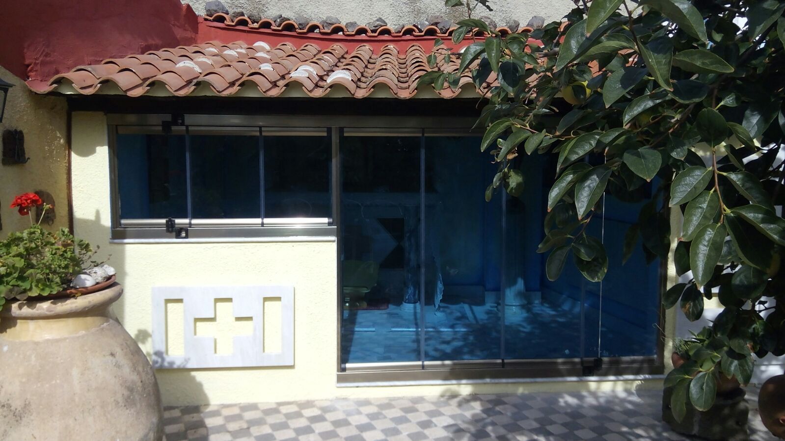 Porta scorrevole in vetro blu, veranda piastrellata, piante in vaso e tetto con tegole rosse. Luce del sole e ombre.