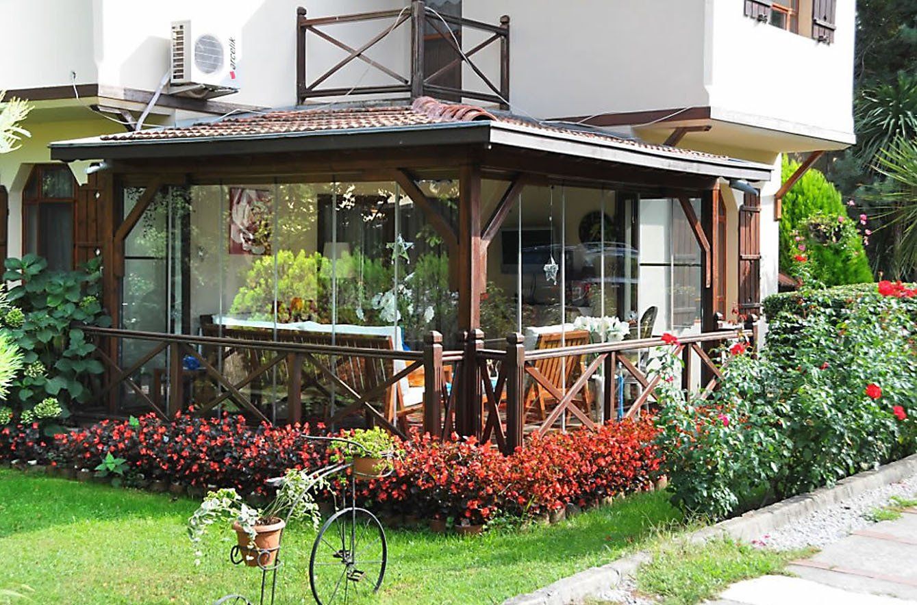Una veranda con struttura in legno e pareti in vetro, circondata da un giardino con fiori rossi ed erba verde.