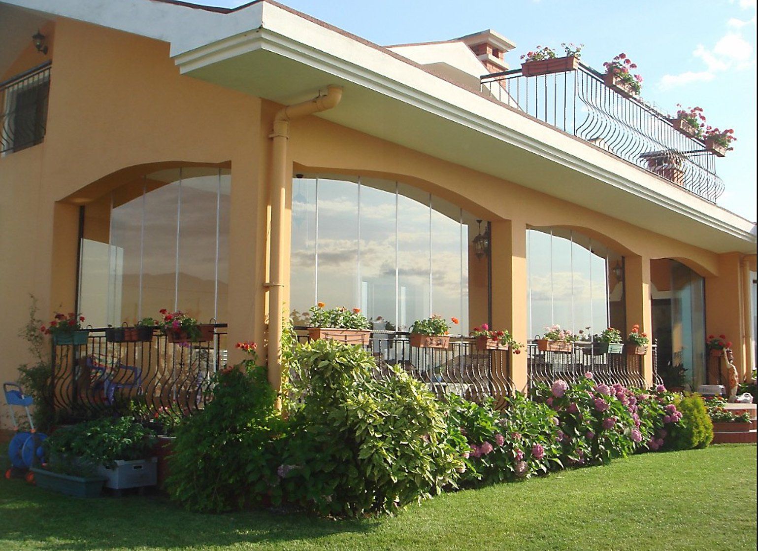 Edificio beige con ampie vetrate, fiori e un balcone sul tetto. Cielo coperto riflesso.