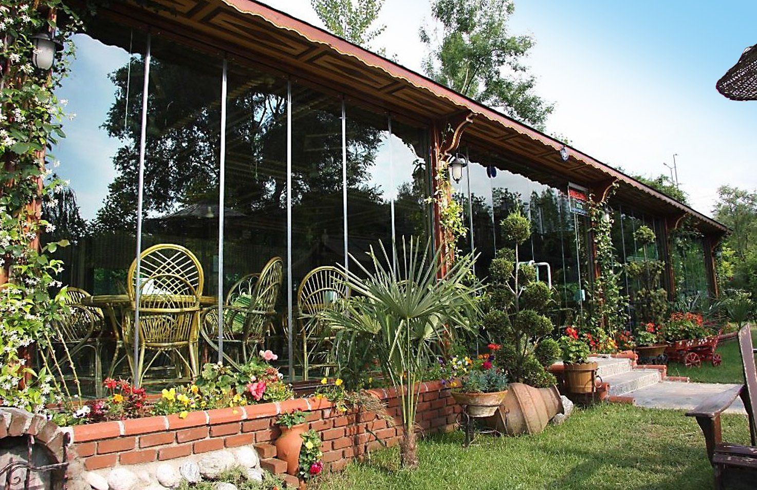 Zona pranzo all'aperto con pareti in vetro, circondata da piante e fiori.