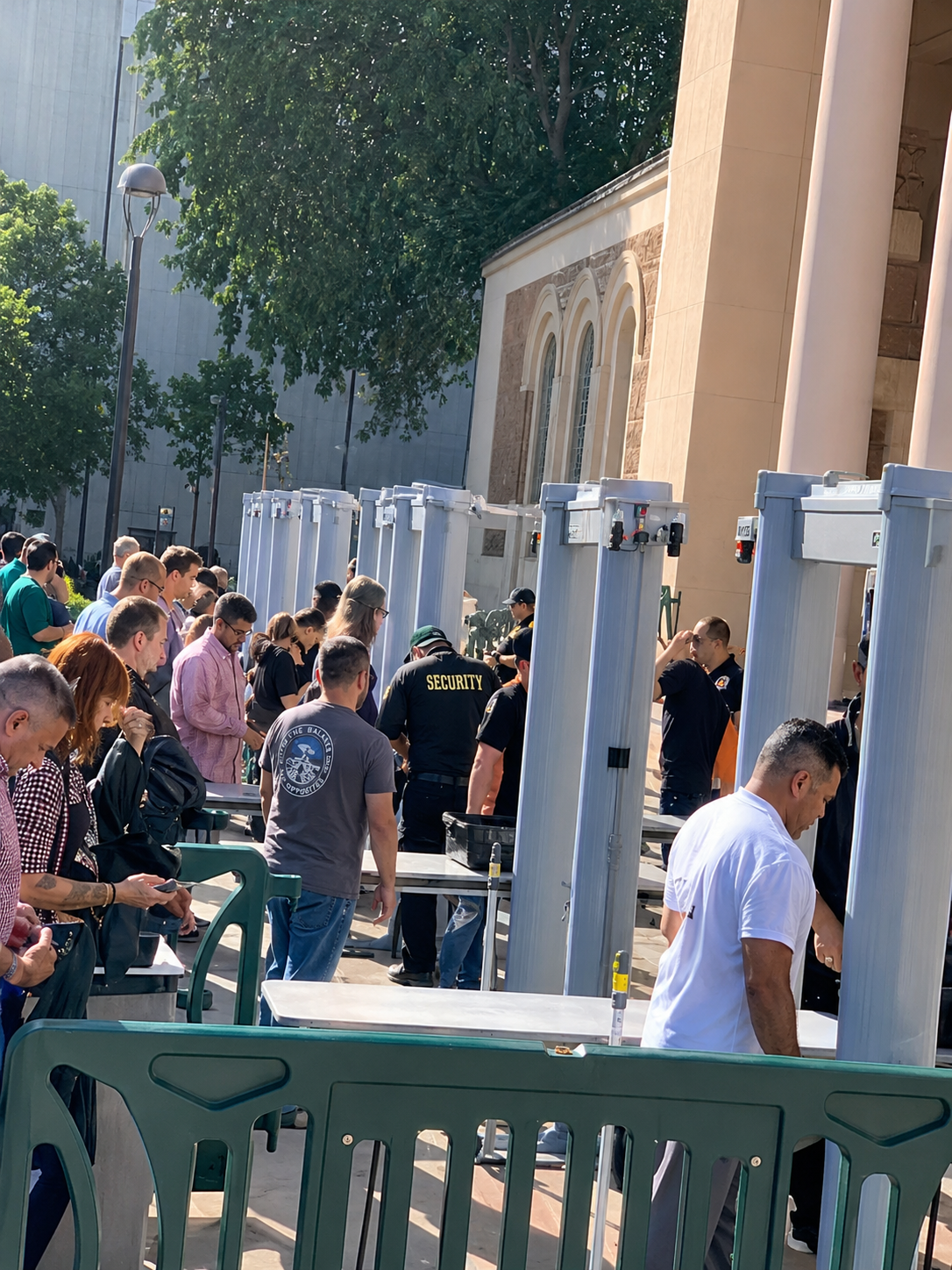 People queuing outside a building beside blue-and-white booths and green barriers in bright sunlight