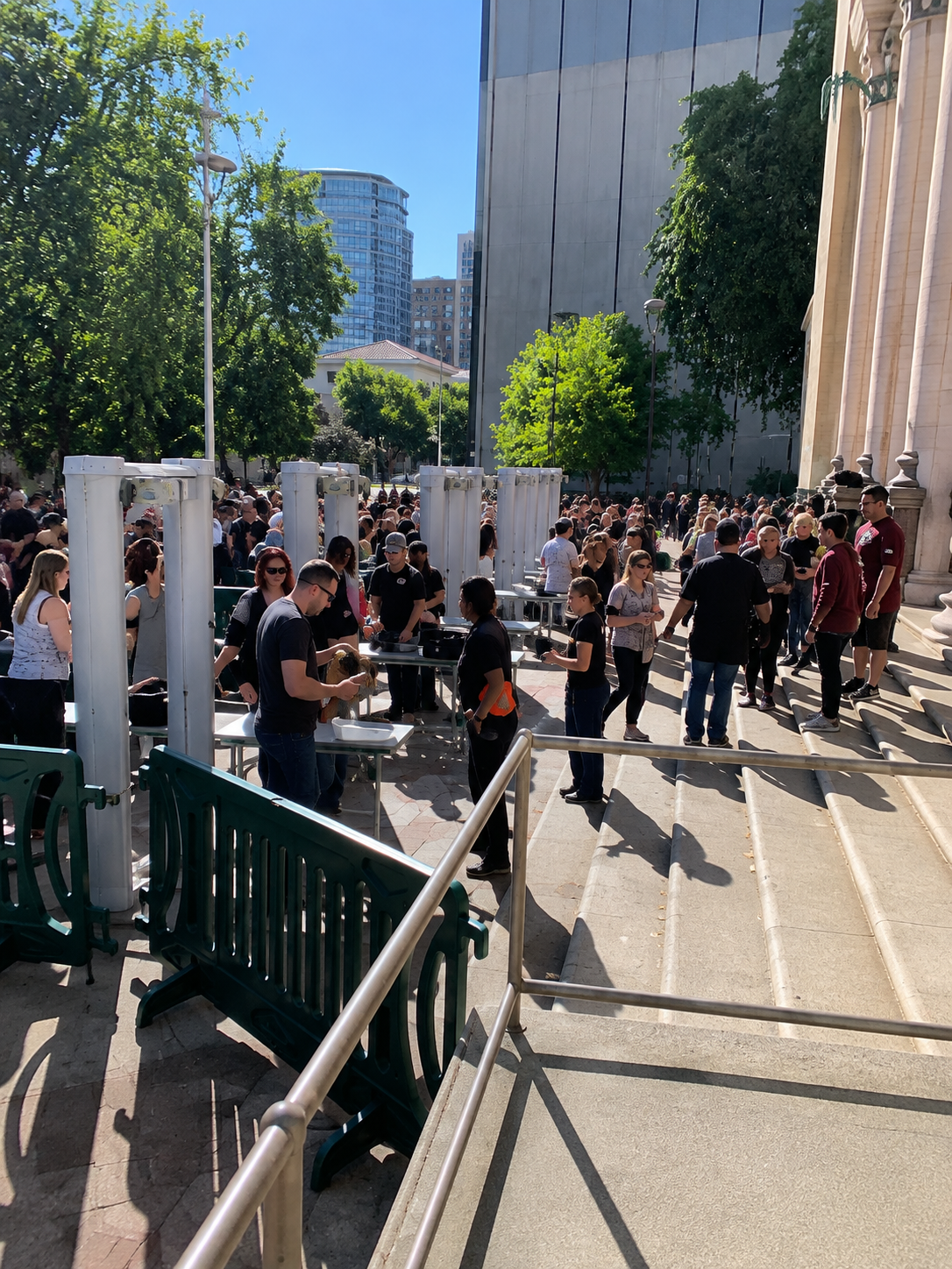 Crowd gathered outside a modern building on a sunny day, with steps, trees, and white columns visible.