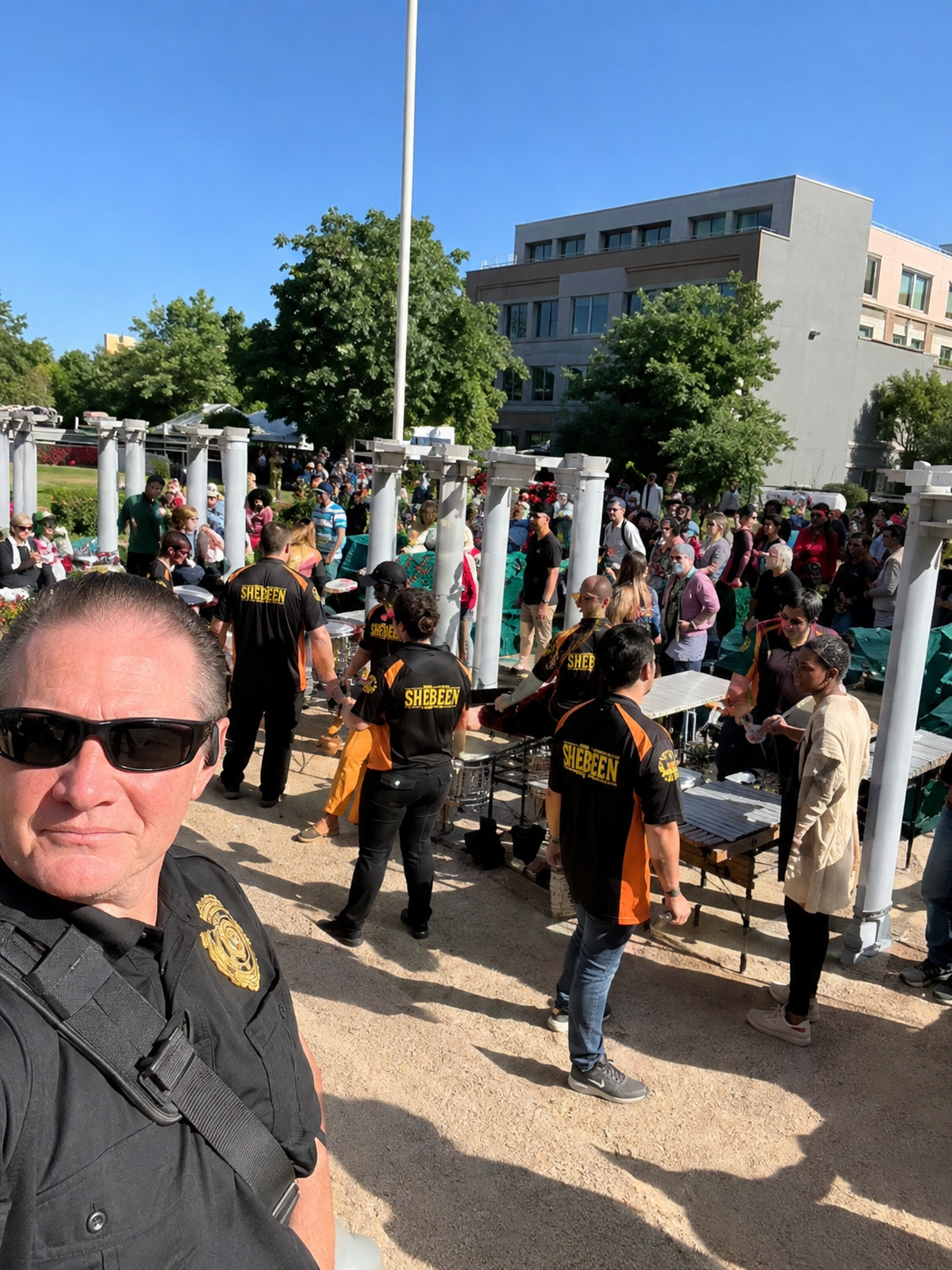 Crowd gathered outdoors in a plaza, with people in colorful shirts and a man taking a selfie in the foreground.