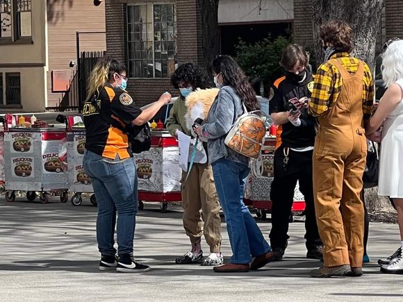 People standing and talking outdoors beside a row of food carts, holding drinks and plates.