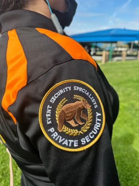 Security guard in black-and-orange uniform with “Private Security” patch, standing outdoors.