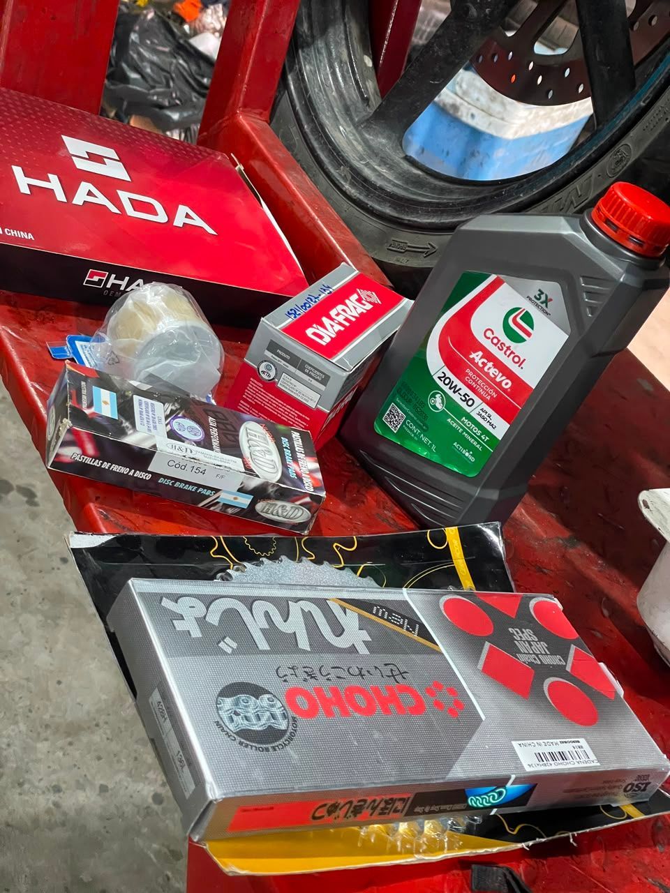 Varias baterías de coche y contenedores apilados sobre una superficie roja, incluyendo una caja gris y una etiqueta de batería verde.