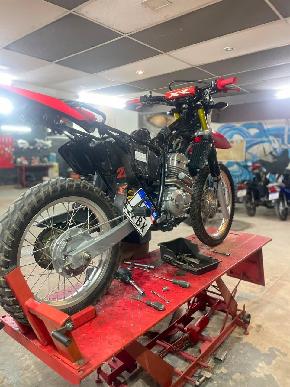 Motocicleta todoterreno negra sobre un elevador rojo en un taller, con herramientas esparcidas cerca.
