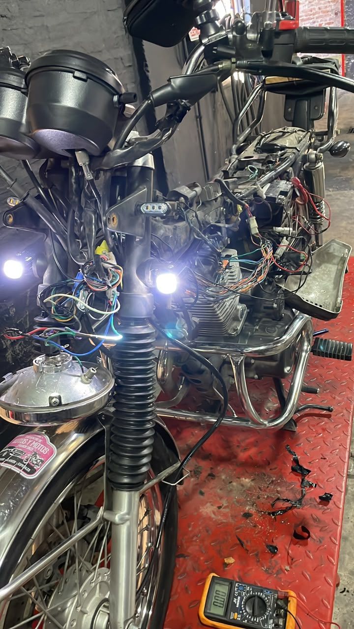 Motocicleta dañada con cables expuestos y faros encendidos sobre un elevador de taller, rodeada de herramientas y escombros.