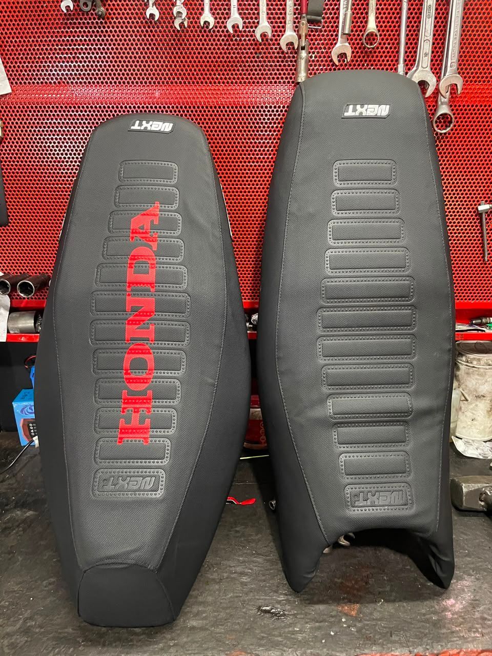 Dos asientos de motocicleta Honda negros con el logotipo rojo de Honda, expuestos uno al lado del otro sobre un fondo de panel perforado rojo.