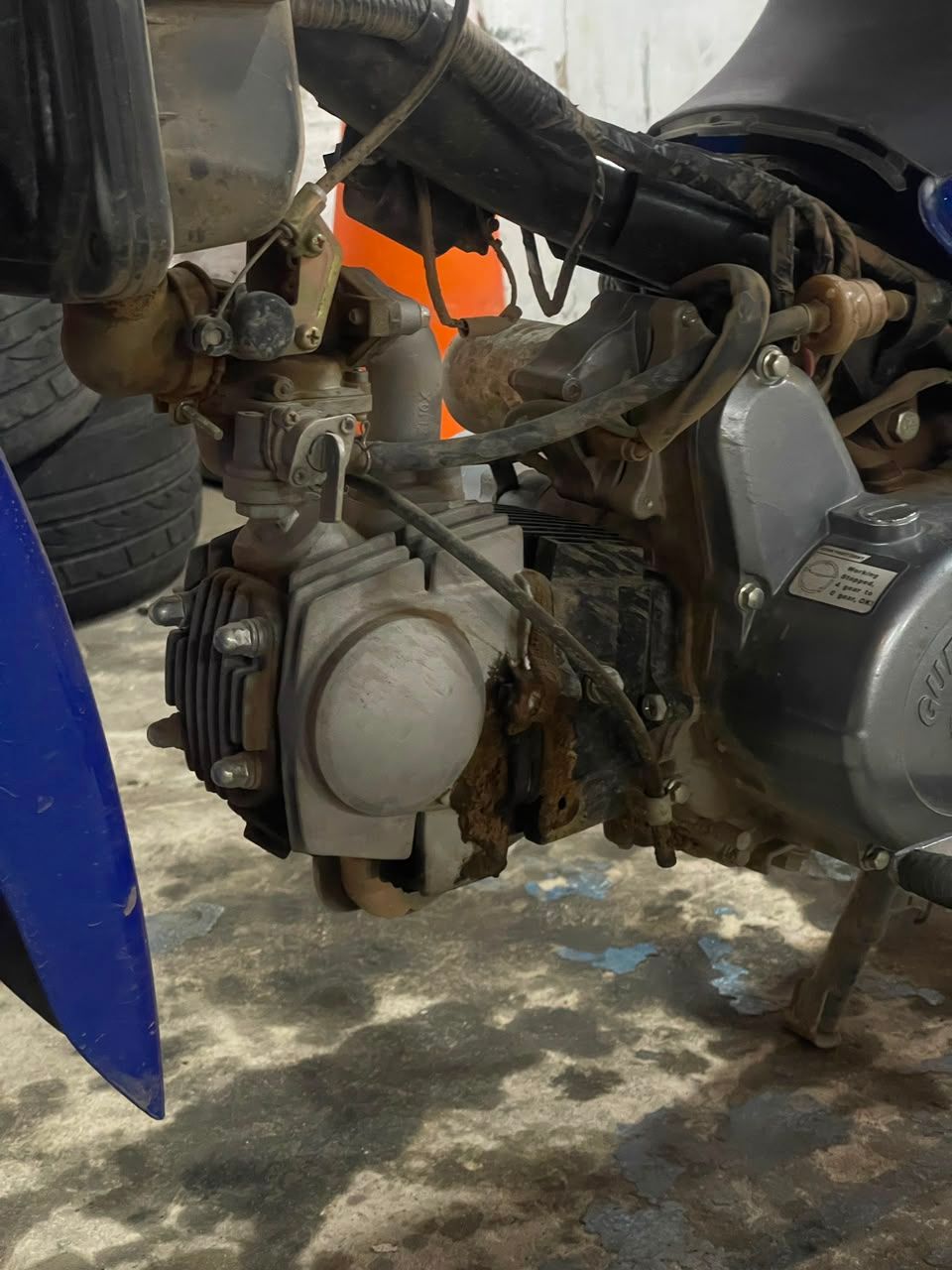 Primer plano del motor y el chasis de una moto de cross con detalles en naranja en un garaje o taller.