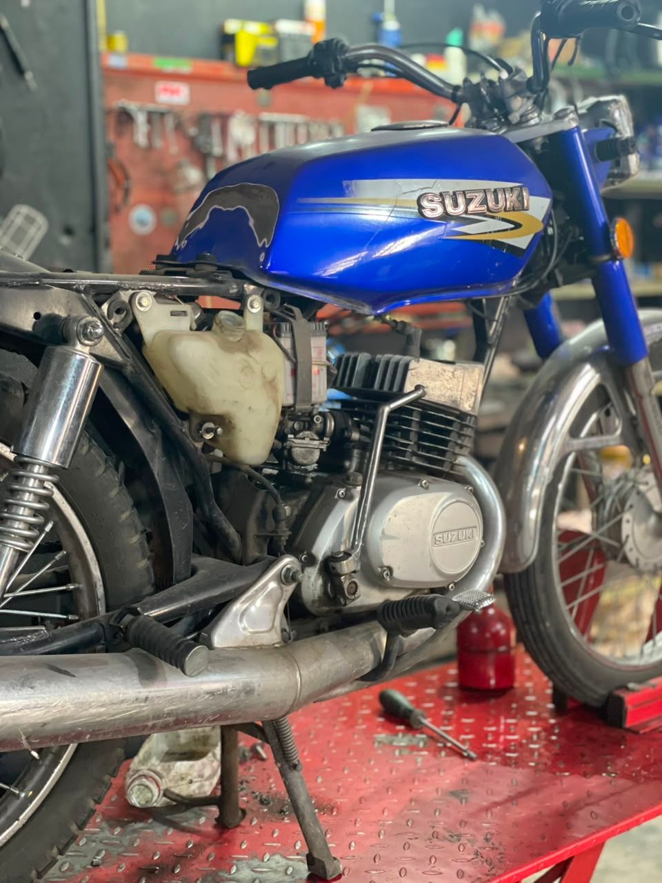 Motocicleta Suzuki azul sobre un banco de trabajo rojo en un garaje, mostrada en una vista lateral en primer plano.