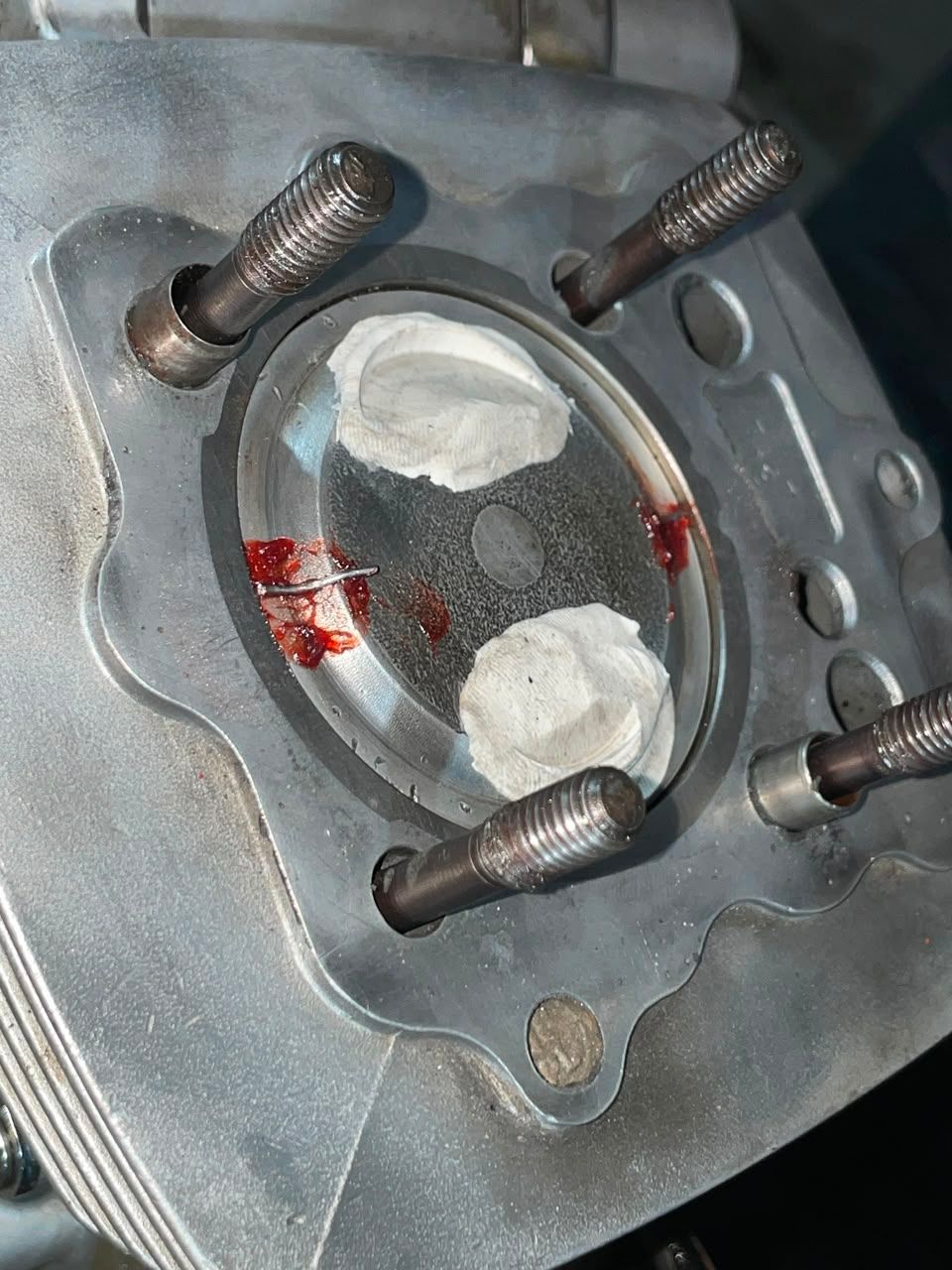 Primer plano de una culata metálica con cuatro tornillos y una abertura cubierta de masilla blanca con residuos rojos.