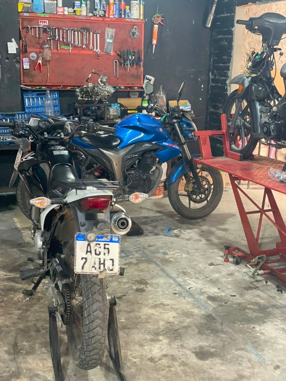 Motocicletas en un garaje desordenado con un armario de herramientas rojo y un banco de trabajo repleto de piezas.