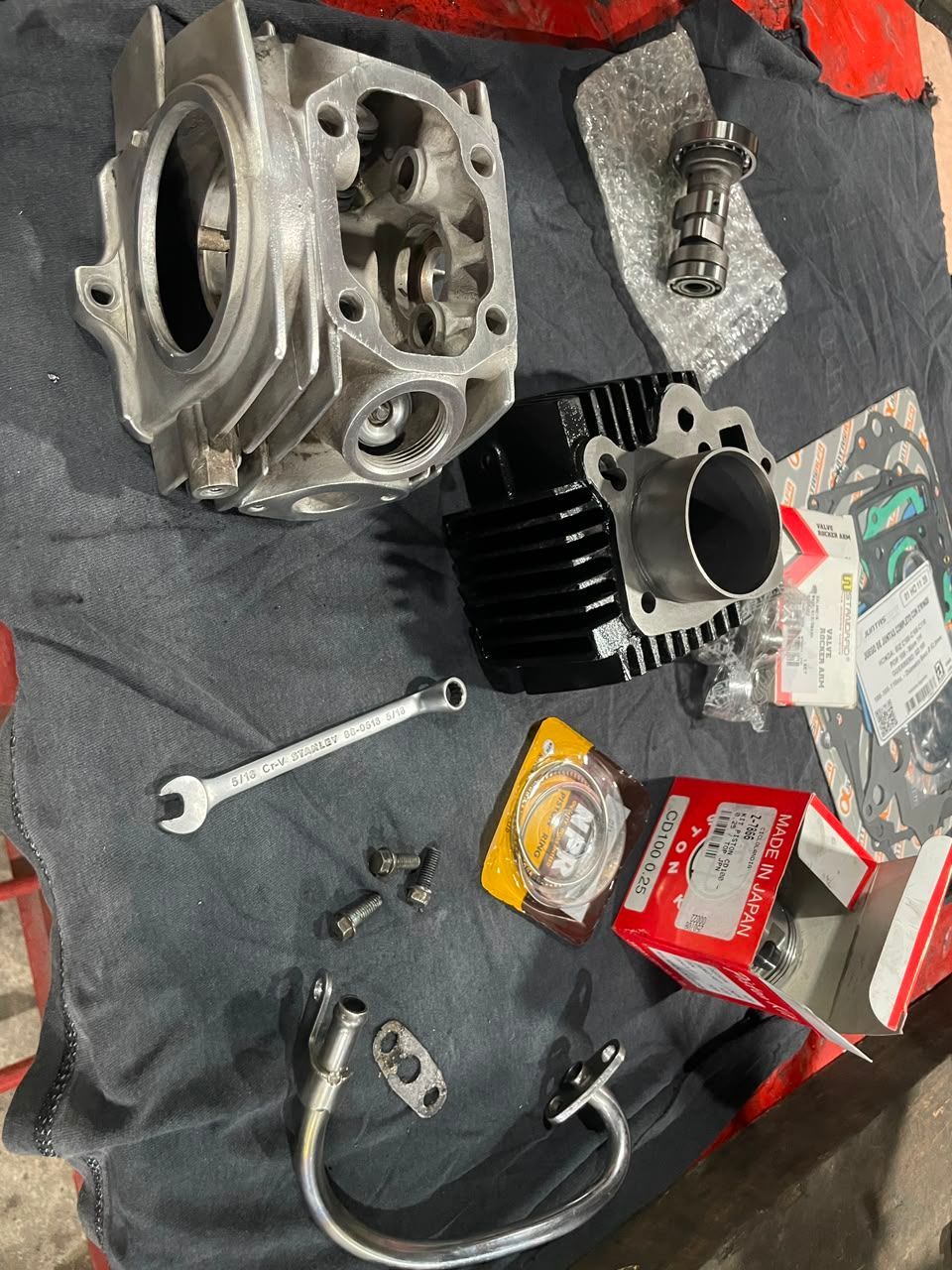 Piezas desmontadas del motor de una motocicleta sobre un paño oscuro, con herramientas y embalaje rojo cerca.