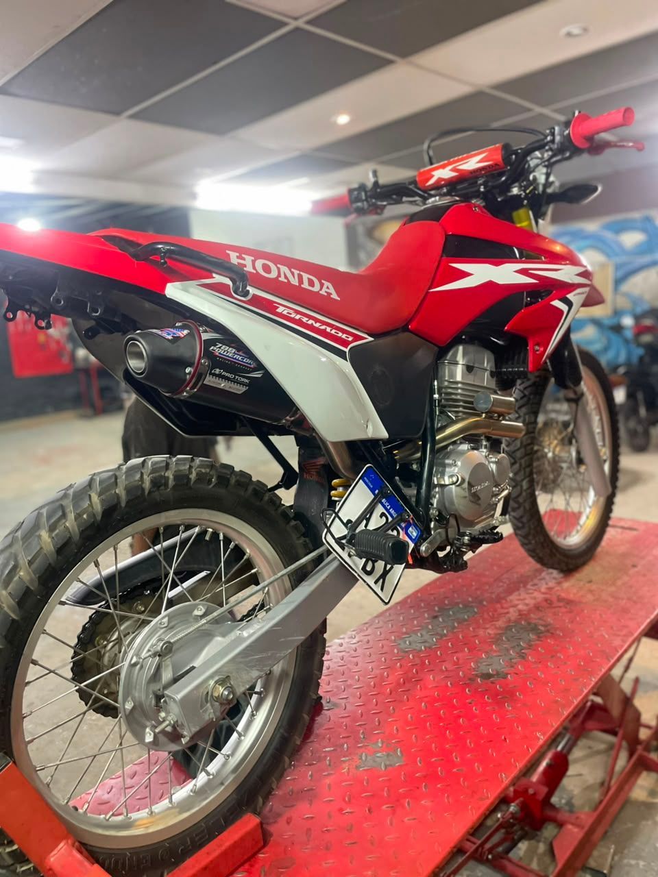 Moto de cross Honda roja sobre una plataforma roja en el interior, vista desde la parte trasera izquierda.