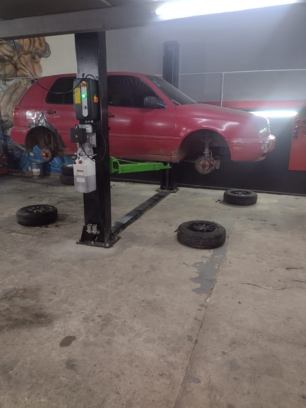 Un coche rojo elevado sobre un elevador de dos columnas en un garaje, con las ruedas desmontadas y los neumáticos en el suelo.