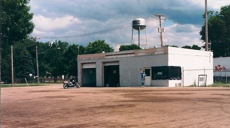 Small white utility building with water towers behind it and a motorcycle parked on a dirt lot. | Boeser Automotive
