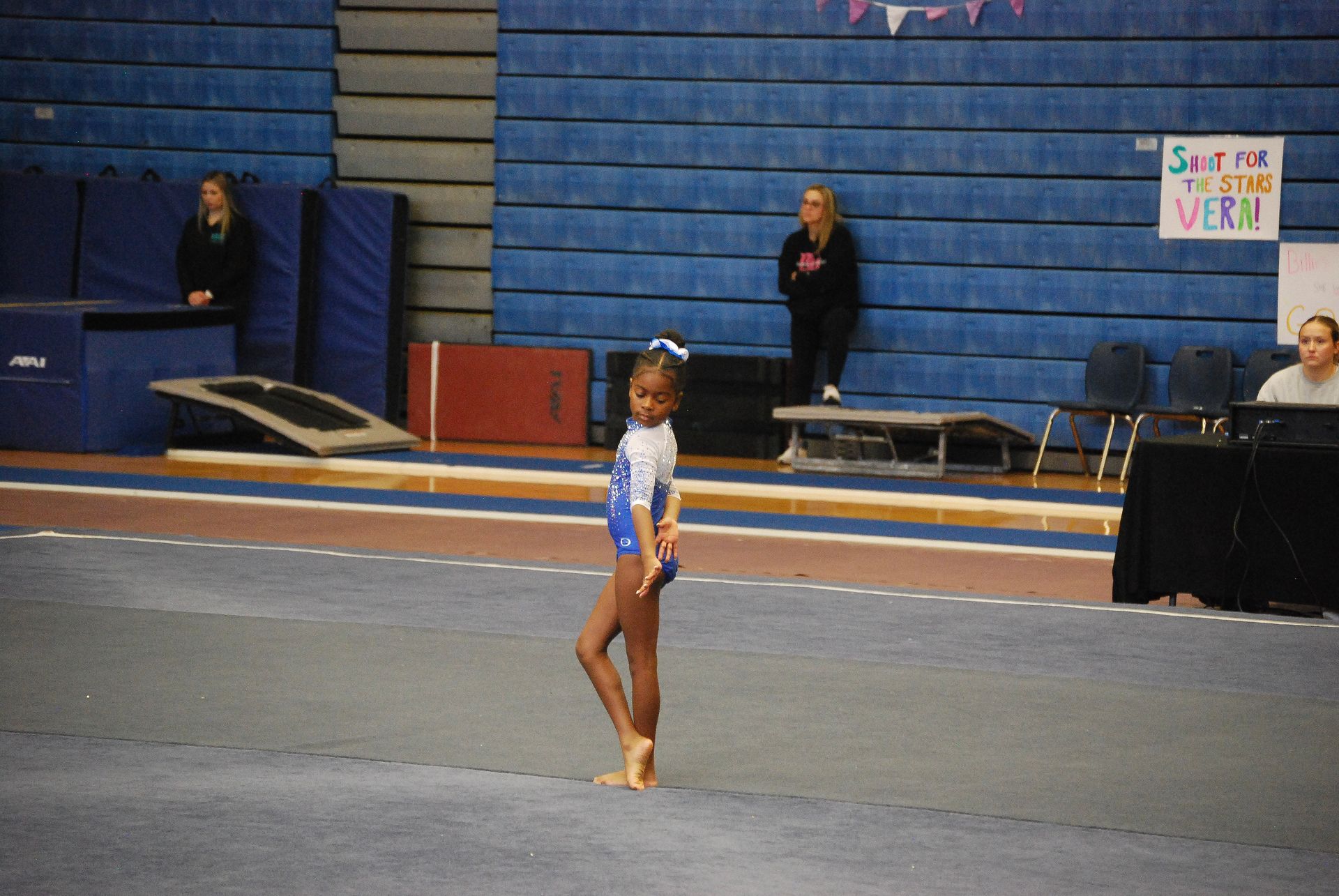 Gymnast in blue leotard on blue mat, arms at side. Gym in background with spectators and equipment.