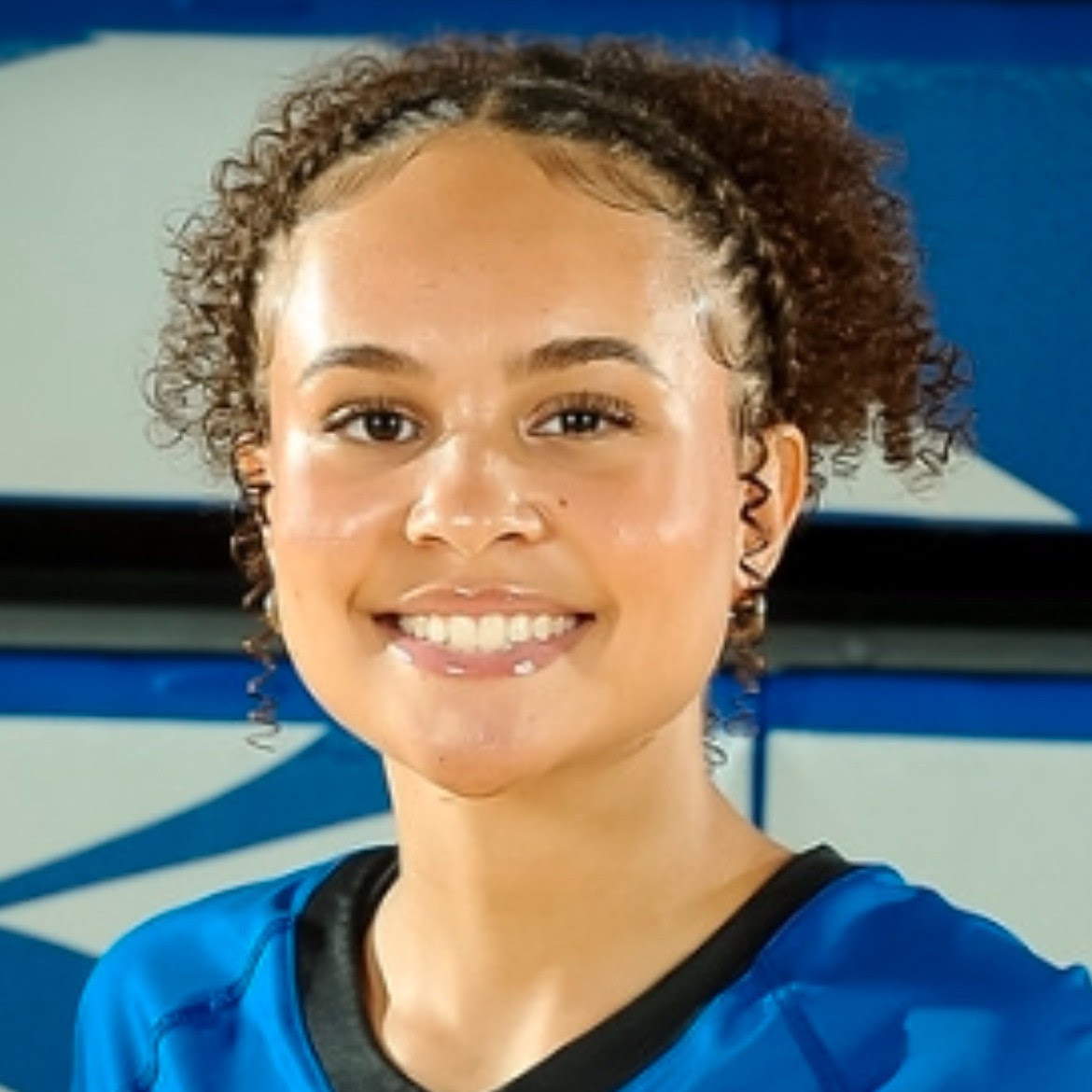 Young woman with curly hair, smiling, in a blue top, in a gymnasium.