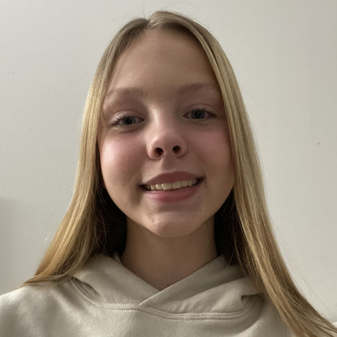 Blonde-haired teen smiles at the camera, wearing a beige hoodie, in front of a white wall.