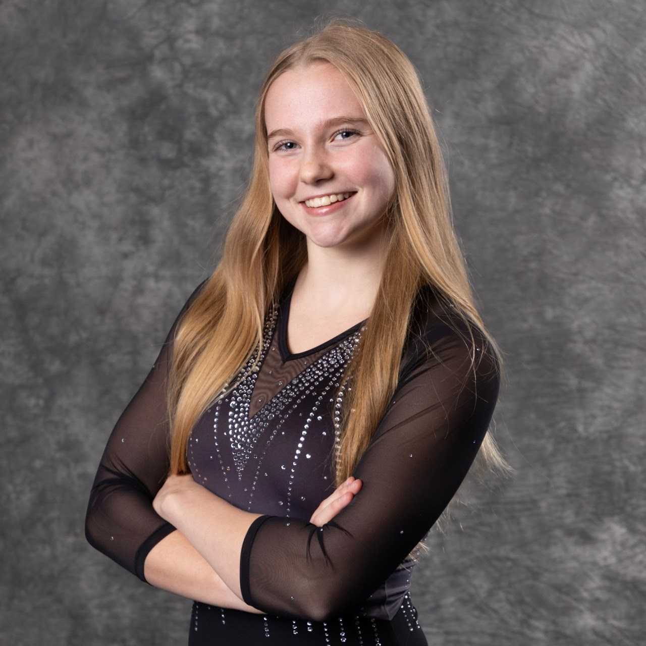 Young blonde woman in gymnastics attire smiling with arms crossed.