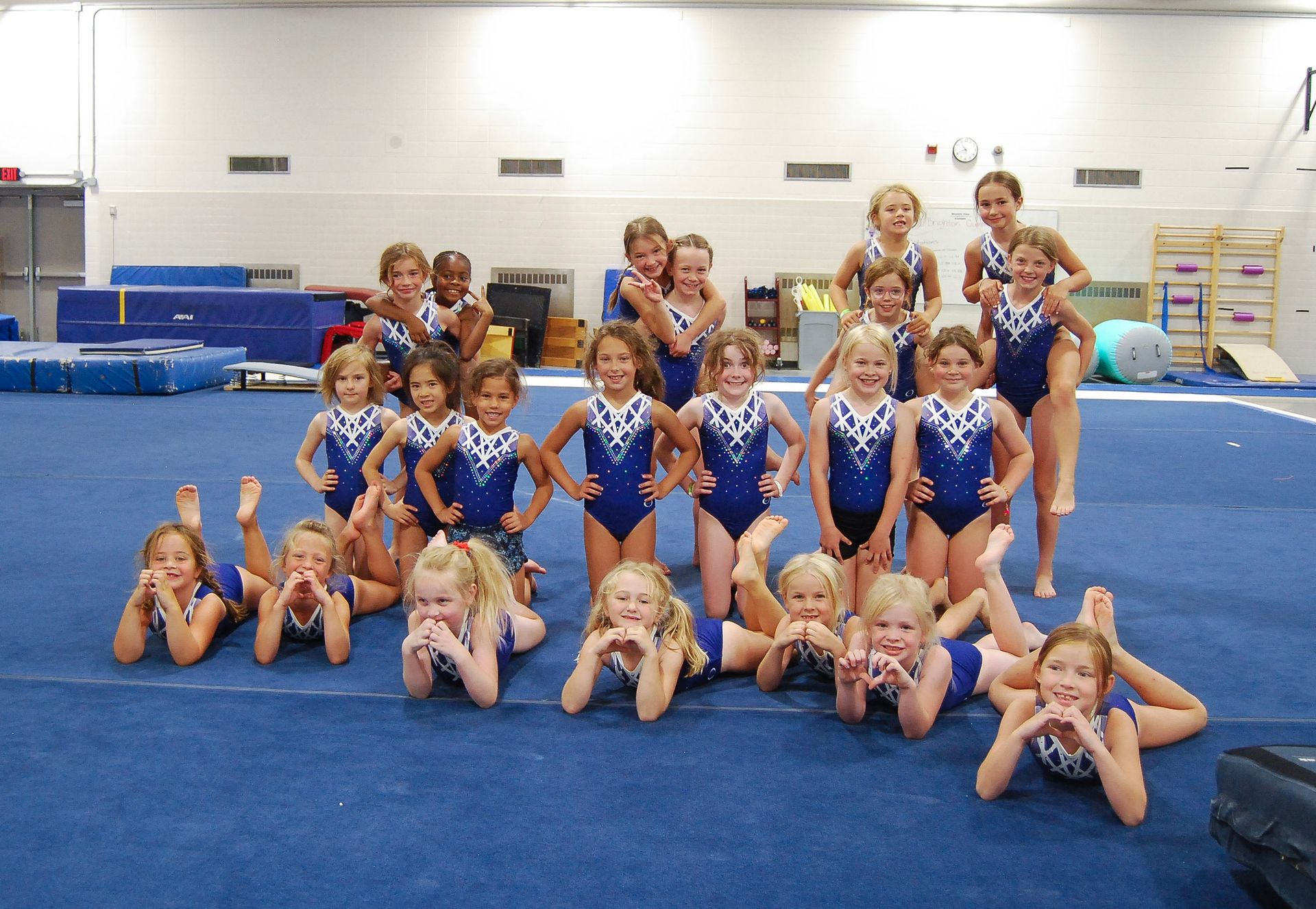 Gymnastics team posing in a gym, wearing blue leotards, some lying on a blue mat.