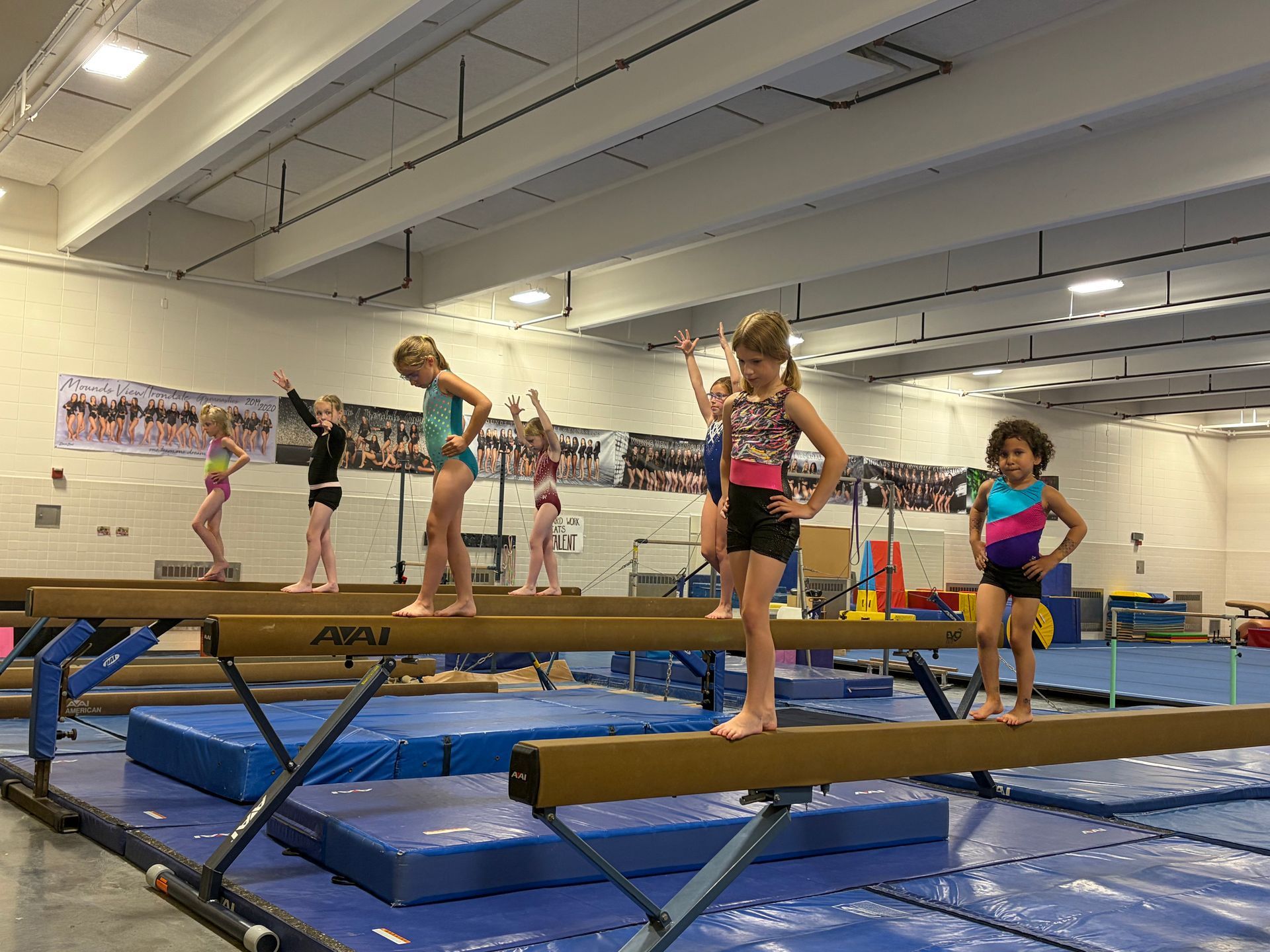 Children practicing balance on beams in a gymnastics gym with blue mats and a mirrored wall