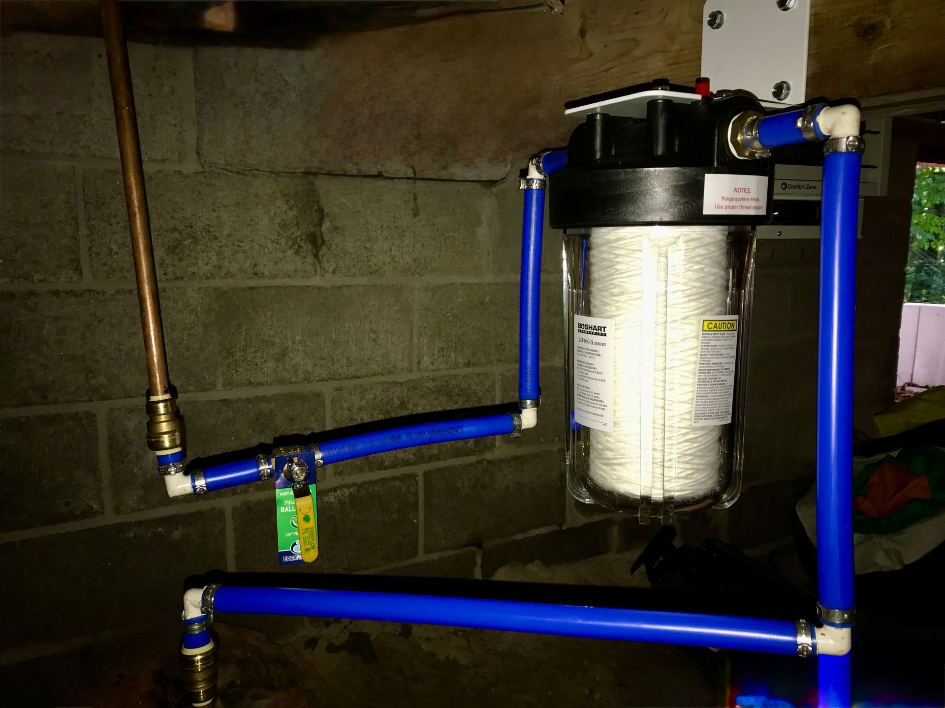Basement water filter setup with blue pipes and a large cylindrical filter mounted on a cinderblock wall