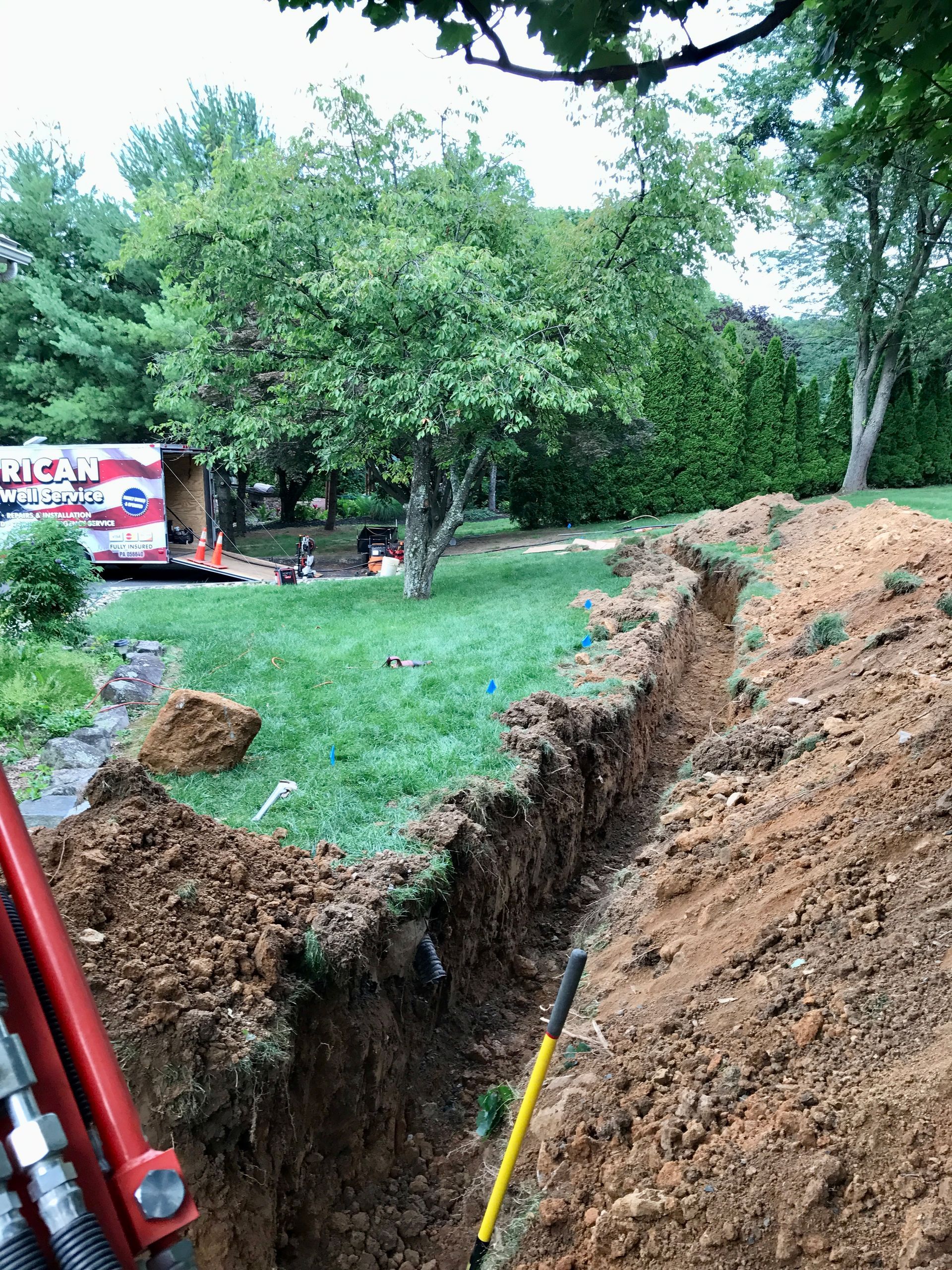 Deep trench cut through a grassy yard with a red excavator arm and trees in the background