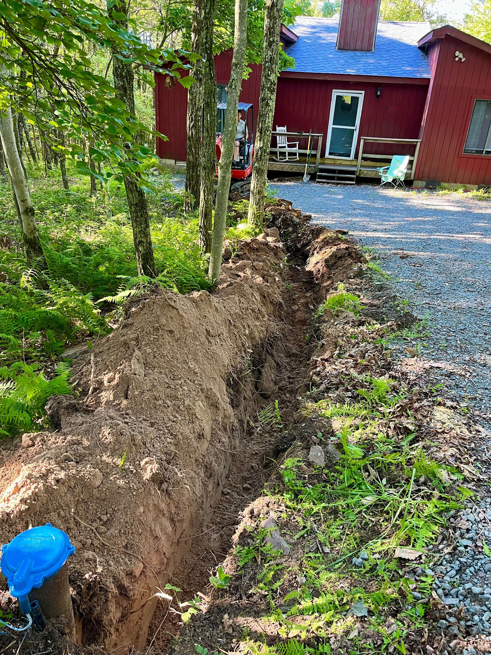 Trench dug beside a red house, with gravel driveway and trees in the foreground.