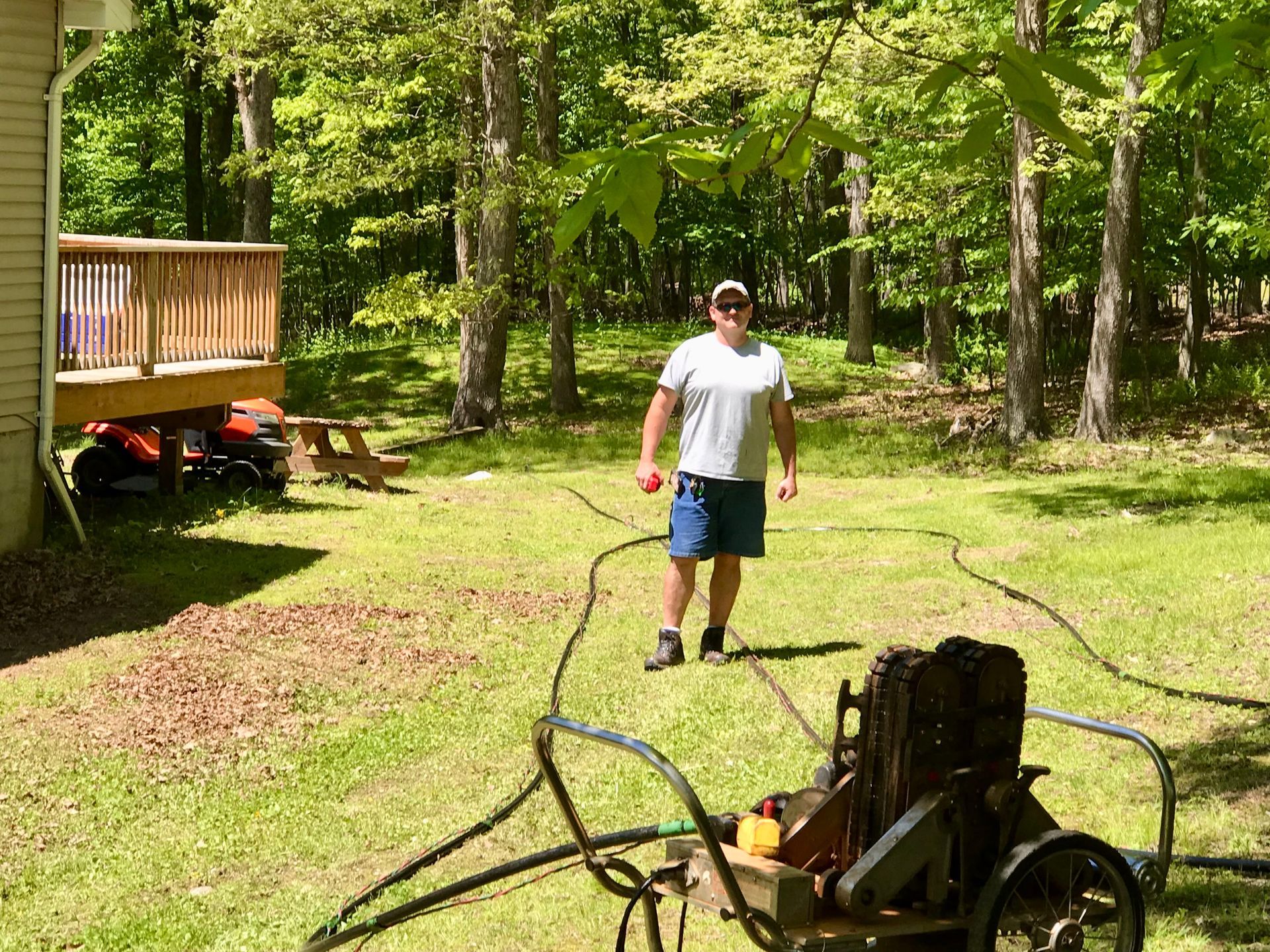 Person standing in a sunny yard near a hose and yard equipment beside a wooden deck