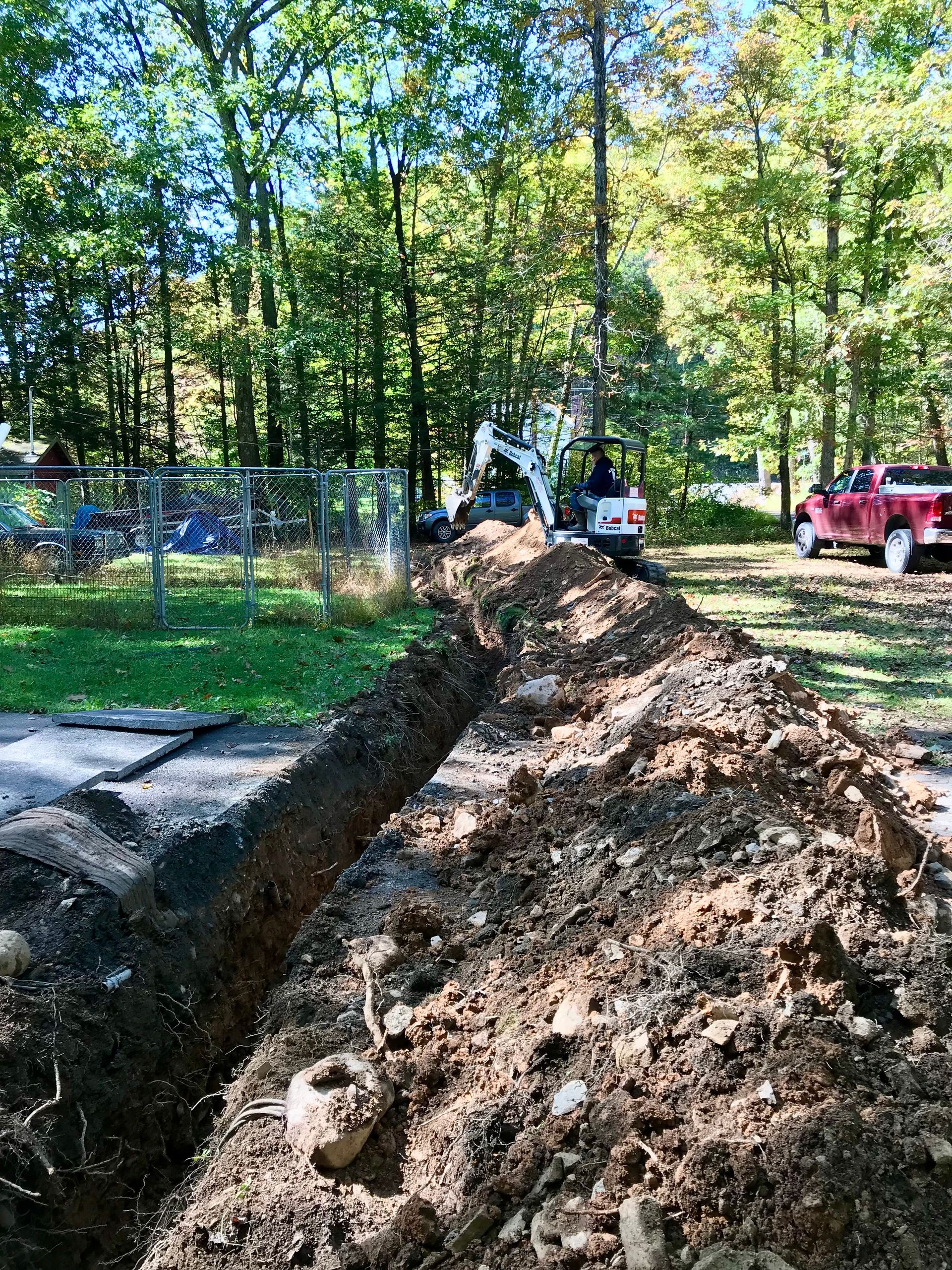 A muddy trench beside a fenced wooded area, with a red pickup truck and equipment in the background.