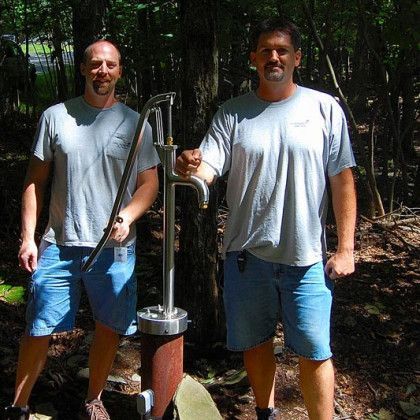 Two men standing outdoors holding a hand water pump in a wooded area