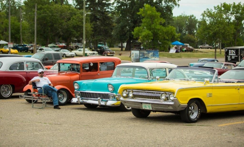 A man is sitting in a chair in front of a row of old cars.
