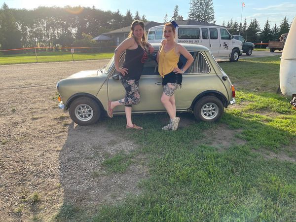 Two women are standing next to a small car in a grassy field.