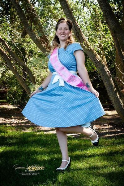 A woman in a blue dress and pink sash is standing in the grass.