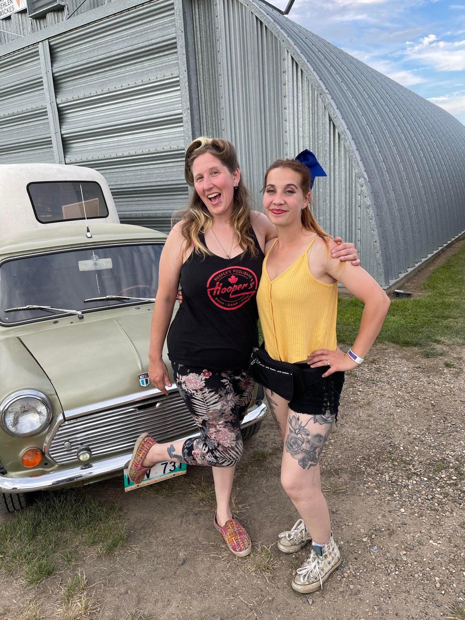 Two women are posing for a picture in front of a car.